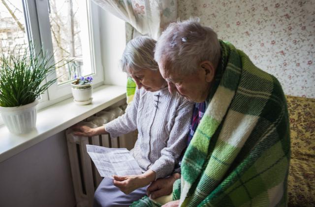 Photograph of an elderly couple which is covered in blankets and is sitting next to a heater studying a bill.