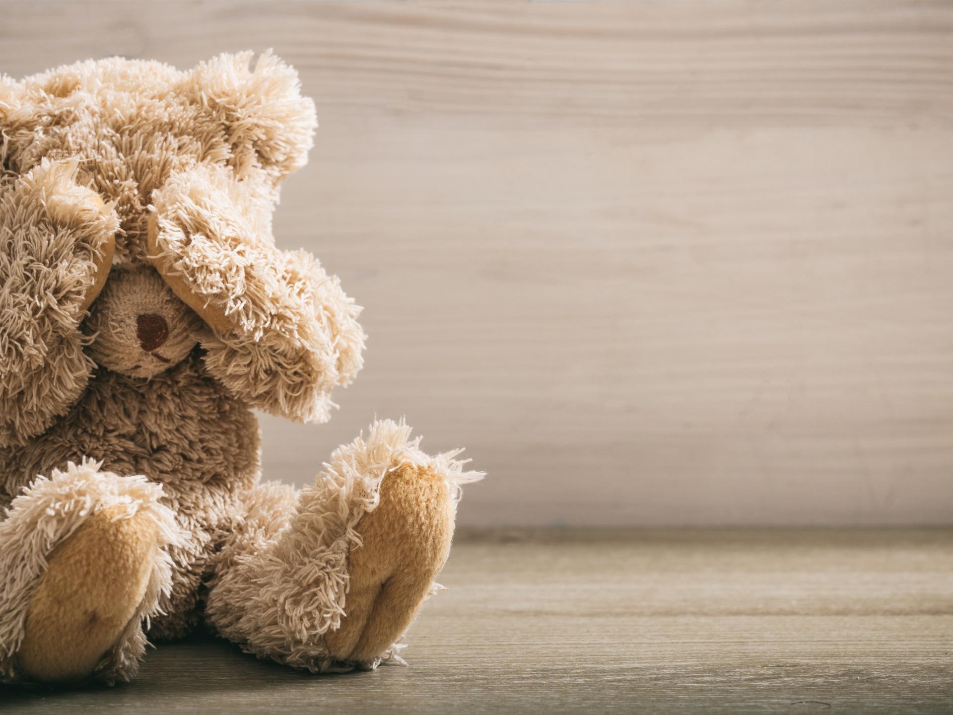 Fluffy beige teddy bear sitting on wooden floor covering its eyes with its paws against a light wood background.