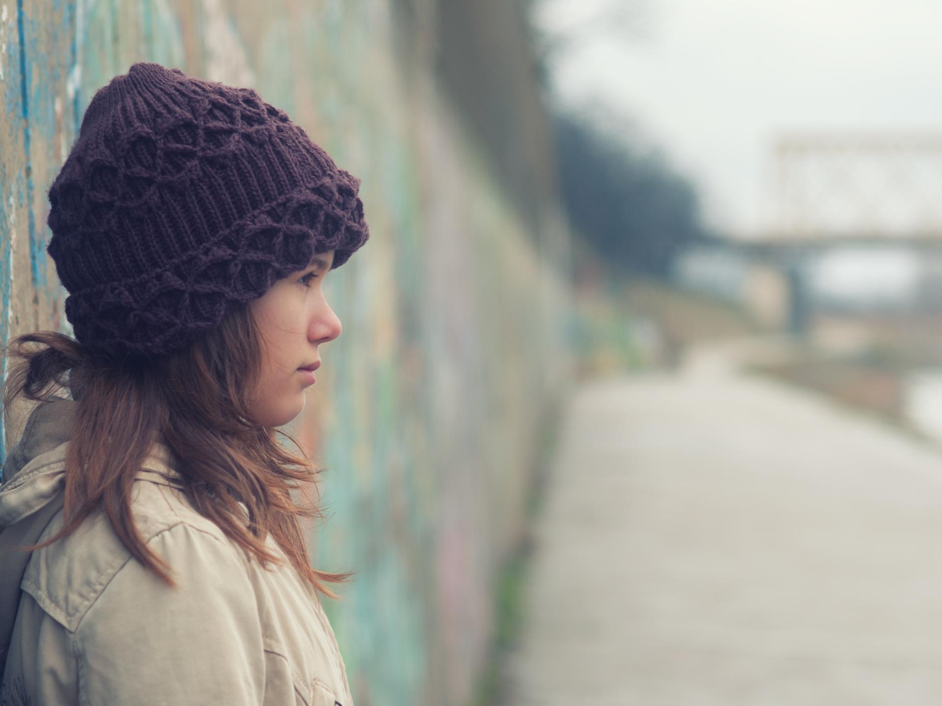 Woman wearing a knitted purple beanie and beige jacket leaning against a graffiti-covered wall by a riverside path.