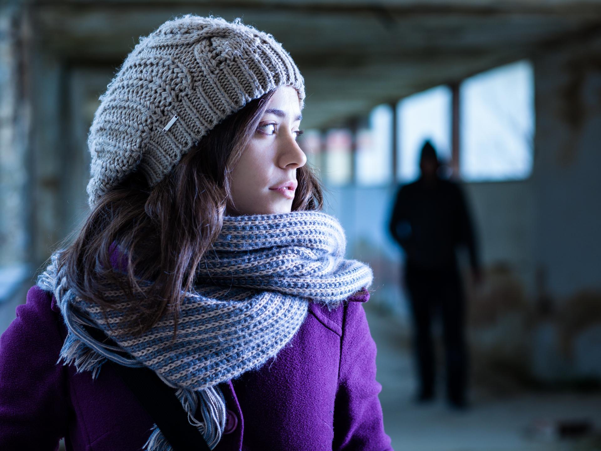 Woman wearing a knitted beige hat and striped scarf stands in a dimly lit abandoned building with a shadowy figure in the background.