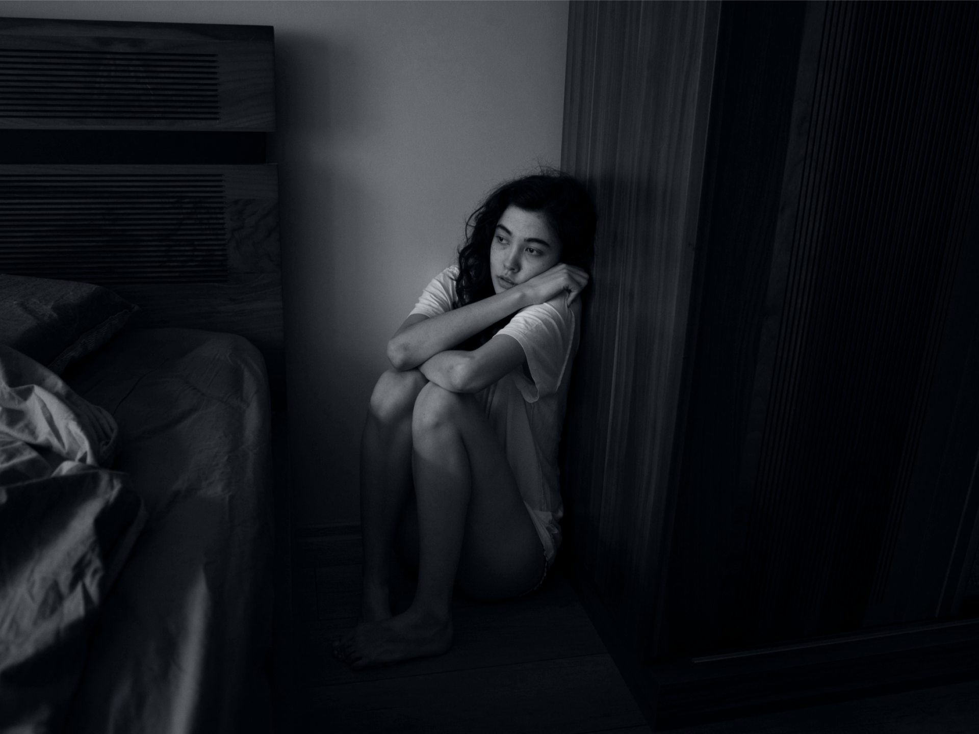 Monochrome image of a woman sitting on the floor, hugging her knees, looking away with a thoughtful expression.